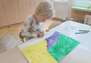 Dziewczynka w białej bluzce w kwiatki siedzi na krzesełku, w dłoni trzyma niebieski mazak i rysuje po śladzie.