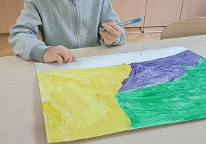 Chłopiec w szarej bluzie siedzi na krzesełku, w dłoni trzyma niebieski mazak i rysuje po śladzie.