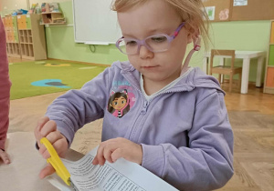 Dziewczynka w fioletowej bluzie i okularach siedzi na krzesełku, w dłoni trzyma żółte nożyczki i wycina kartę.