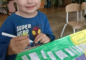 Chłopiec w niebieskiej bluzce siedzi przy stoliku i pisze po śladzie niebieskim mazakiem.