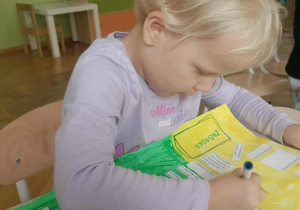 Dziewczynka w blond włosach siedzi przy stoliku i pisze po śladzie niebieskim mazakiem.