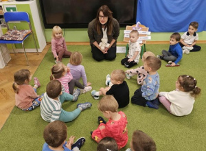 Dzieci siedzą na zielonym dywanie razem z panią pedagog. Pani trzyma w dłoni jasnego misia.
