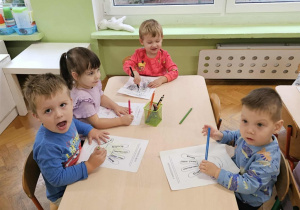 Czwórka dzieci siedzi przy stoliku i koloruje kontur dłoni. Z tyłu widać okna.