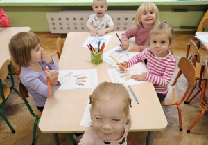 Piątka dzieci siedzi przy stoliku i koloruje kontur dłoni. Z tyłu widać okna.