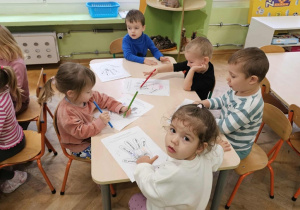 Piątka dzieci siedzi przy stoliku i koloruje kontur dłoni. Z tyłu widać okna.