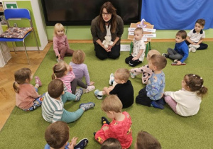 Dzieci siedzą na zielonym dywanie razem z panią pedagog. Pani trzyma w dłoni jasnego misia.