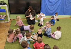 Dzieci siedzą na zielonym dywanie razem z panią pedagog. Pani trzyma w dłoni jasnego misia.