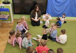 Dzieci siedzą na zielonym dywanie razem z panią pedagog. Pani trzyma w dłoni jasnego misia.