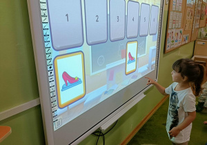 Dziewczynka w białej bluzce dotyka ekranu. Na ekranie widać dwa buty.