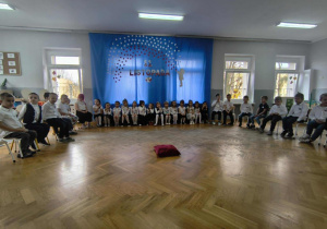 Dzieci siedzą w półkolu na krzesłach. Z tyłu wisi dekoracja na niebieskim materiale.