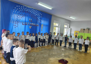 Dzieci stoją w półkolu. W tle wisi dekoracja na niebieskim materiale,