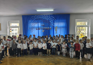 Dzieci stoją na tle niebieskiej dekoracji. Część dzieci trzyma w dłoniach flagi.