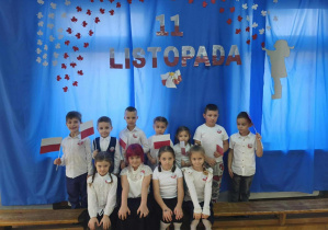 11 dzieci stoi na tle niebieskiej dekoracji. W dłoniach trzymają flagi.