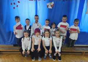 11 dzieci stoi na tle niebieskiej dekoracji. W dłoniach trzymają flagi.