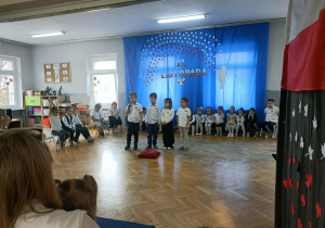 Czwórka dzieci stoi na środku i recytuje wiersz. Dzieci stoją z tyłu w półkolu.