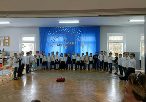 Dzieci siedzą w półkolu na krzesłach. Z tyłu wisi dekoracja na niebieskim materiale.