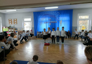 Piątka dzieci stoi na środku i recytuje wiersz. Dzieci stoją z tyłu w półkolu.