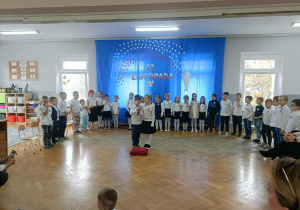 Dwójka dzieci stoi na środku i recytuje wiersz. Dzieci stoją z tyłu w półkolu.