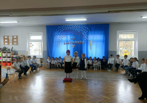 Trójka dzieci stoi na środku i recytuje wiersz. Dzieci stoją z tyłu w półkolu.