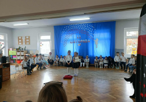 Dwie dziewczynki stoją na środku i recytują wiersz. Dzieci stoją z tyłu w półkolu.