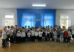 Dzieci stoją na tle niebieskiej dekoracji. Część dzieci trzyma w dłoniach flagi.