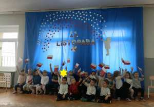 Dzieci stoją i siedzą na tle niebieskiej dekoracji. Część dzieci trzyma w dłoniach flagi.