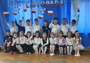 Dzieci stoją i siedzą na tle niebieskiej dekoracji. Część dzieci trzyma w dłoniach flagi.