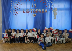 Dzieci stoją i siedzą na tle niebieskiej dekoracji. Część dzieci trzyma w dłoniach flagi.