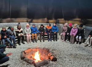 Grupa dzieci siedzi na ławkach wokół ogniska.