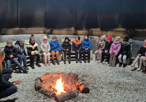 Grupa dzieci siedzi na ławkach wokół ogniska.