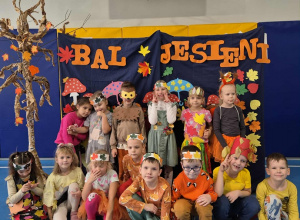 Dzieci w przebraniach stoją na tle dekoracji z napisem Bal Jesieni. Po lewej stronie stoi papierowe drzewo.