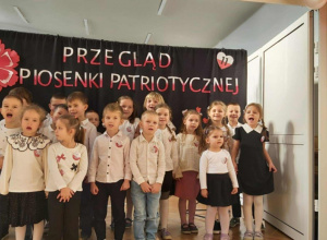 Grupa dzieci stoi na tle niebieskiej dekoracji. Na niebieskim materiale jest napis "Przegląd Piosenki Patriotycznej"