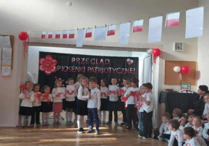 Dwójka dzieci recytuje wiersz. Grupa dzieci stoi z tyłu, na tle dekoracji na niebieskim materiale.