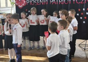 Dwójka dzieci recytuje wiersz. Grupa dzieci stoi z tyłu, na tle dekoracji na niebieskim materiale.