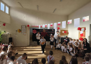 Grupa dzieci siedzi wokół sceny. Przy suficie wiszą flagi. Na dekoracji jest napis "Przegląd Piosenki Patriotycznej"