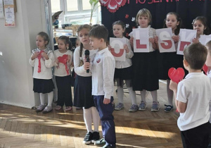 Dwójka dzieci recytuje wiersz. Grupa dzieci stoi z tyłu, na tle dekoracji na niebieskim materiale.
