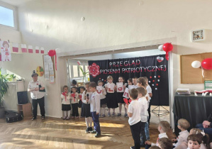 Dwójka dzieci recytuje wiersz. Grupa dzieci stoi z tyłu, na tle dekoracji na niebieskim materiale.