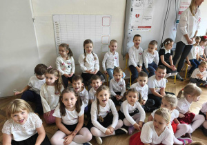 Grupa dzieci siedzi na parkiecie w eleganckich strojach.