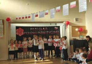 Dwójka dzieci recytuje wiersz. Grupa dzieci stoi z tyłu, na tle dekoracji na niebieskim materiale.