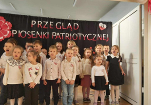 Grupa dzieci stoi na tle niebieskiej dekoracji. Na niebieskim materiale jest napis "Przegląd Piosenki Patriotycznej"