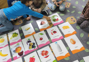 Na dywanie leżą plansze z jedzeniem. Dzieci siedzą wokół. Chłopiec w niebieskiej bluzie pokazuje palcem na plansze.