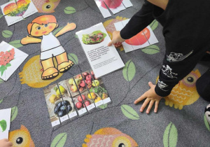 Na dywanie leżą ułożone puzzle. Chłopiec pokazuje palcem plansze ze zdrową kanapką.