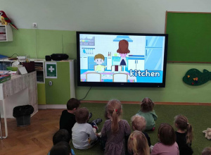 Dzieci siedzą na zielonym dywanie i oglądają film edukacyjny.