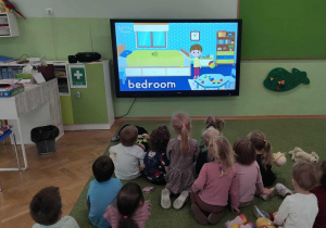 Dzieci siedzą na zielonym dywanie i oglądają film edukacyjny.