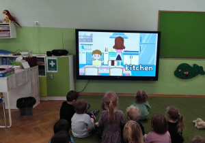 Dzieci siedzą na zielonym dywanie i oglądają film edukacyjny.