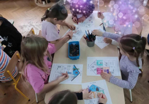 Dzieci siedzą przy stolikach i kolorują kolorowankę przedstawiającą dom.