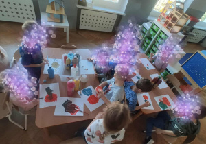 Dzieci siedzą przy stolikach i malują farbami.