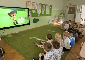 Dzieci siedzą na zielonym dywanie i oglądają bajkę "Kot w butach"