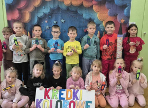 Dzieci stoją na tle kolorowej dekoracji. W dłoniach trzymają papierowe kredki.