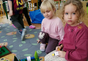 Dwie dziewczynki budują z klocków. W tle widać okna i dzieci.